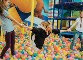 mom in a ball pit with a toddler on her back and two older daughters pulling on each of her arms